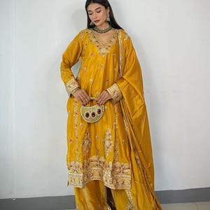 May include: Mustard yellow embroidered salwar kameez set. The outfit features a long-sleeved tunic with intricate embroidery around the neckline, cuffs, and hem. Matching wide-leg trousers and a draped scarf complete the ensemble. A small, decorative clutch is held.