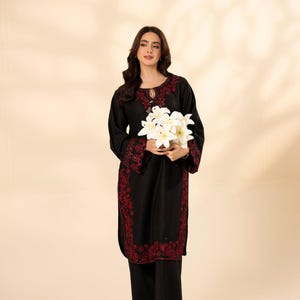 May include: A black ensemble featuring a long-sleeved tunic and matching pants, adorned with intricate red embroidery along the neckline, cuffs, and hem. The outfit is paired with a bouquet of white lilies, creating a striking contrast against the dark fabric.