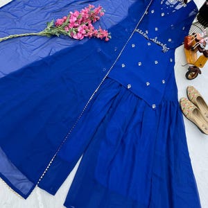 May include: A vibrant blue Sharara suit set. The ensemble includes a top with intricate silver embellishments, matching wide-leg pants, and a sheer dupatta. Pink floral accents and embroidered shoes complement the outfit. The outfit is displayed on a white surface.