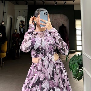 May include: A long-sleeved, purple and white floral print dress with a high neckline and a fitted bodice. The dress has a flowing skirt with a side slit and a silver chain detail.