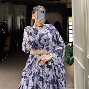 May include: A woman wearing a purple and white floral print dress with a high neck and long sleeves. The dress has a ruffled hem and is made of a sheer fabric.
