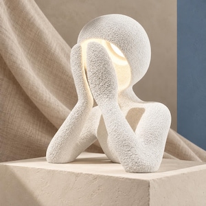 Matte textured white sculpture lamp glowing soft warm white, resting on a beige block with draped beige fabric and a blue panel behind it.