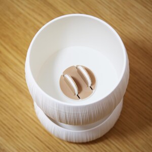 Top-down view into the textured matte white bowl of a self watering planter, showing a round wicking core with a cotton wick at the center against a warm amber-brown background.