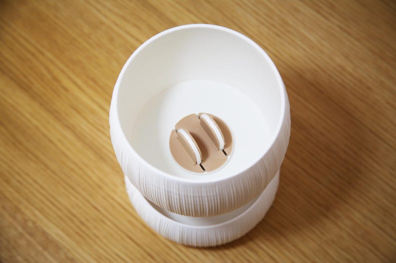 Top-down view into the textured matte white bowl of a self watering planter, showing a round wicking core with a cotton wick at the center against a warm amber-brown background.