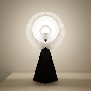 Front view of a small table lamp with a pleated matte white ring shade glowing bright white around the bulb, on a matte black pyramid base against a plain beige wall.