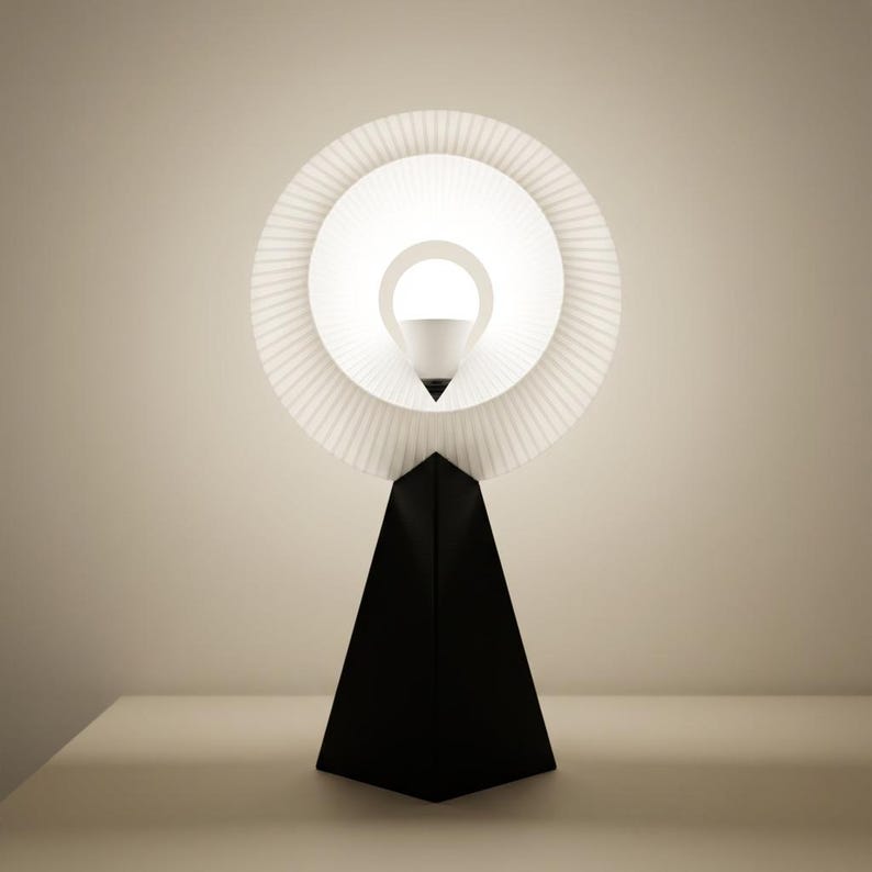 Front view of a small table lamp with a pleated matte white ring shade glowing bright white around the bulb, on a matte black pyramid base against a plain beige wall.