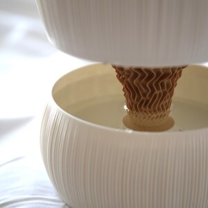 Close-up of the brown cone-shaped core rising from the lower bowl of a textured matte white self watering planter, photographed on soft white fabric.