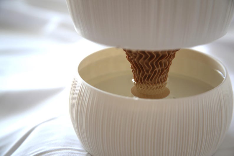 Close-up of the brown cone-shaped core rising from the lower bowl of a textured matte white self watering planter, photographed on soft white fabric.