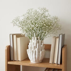 Matte white textured ikebana vase with baby's breath stems on a shelf beside books, showing the vase in a japandi decor setting.