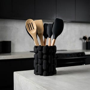 Textured matte black vase holding wooden and black utensils on a white-veined tabletop counter in a dark kitchen.