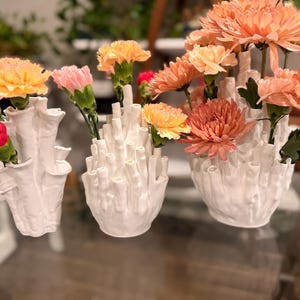 Three white coral vases of different sizes on a glass table, their textured tube openings filled with orange, peach and pink mums and red and pink carnations, with plants in the background.