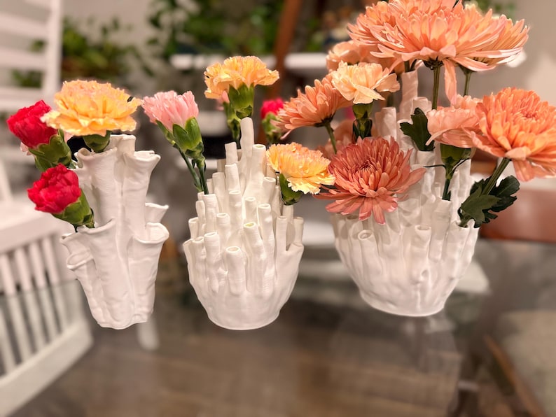 Three white coral vases of different sizes on a glass table, their textured tube openings filled with orange, peach and pink mums and red and pink carnations, with plants in the background.