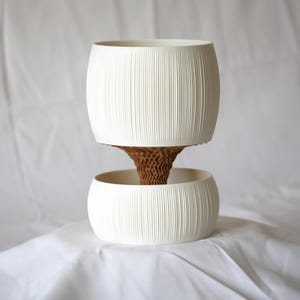 Front view of a cream-white textured matte planter with vertical ribbing, a ribbed pot above a round lower bowl joined by a brown honeycomb-textured core, on white fabric.