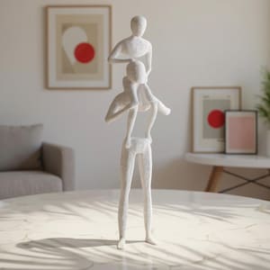 May include: A white sculpture depicting two figures, one atop the other. The bottom figure stands upright, supporting the second figure, who is positioned above. The sculpture is placed on a marble-topped table, with framed artwork in the background.