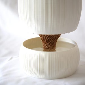 Side view of a textured matte white planter with vertical ribbing, its upper pot and lower bowl joined by a brown cone-shaped core, on white fabric; a self watering pot with a modern look.