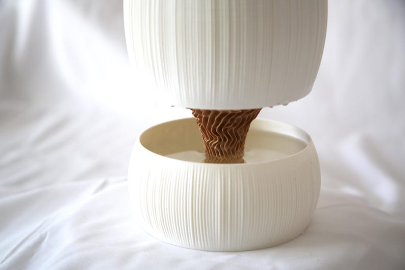 Side view of a textured matte white planter with vertical ribbing, its upper pot and lower bowl joined by a brown cone-shaped core, on white fabric; a self watering pot with a modern look.
