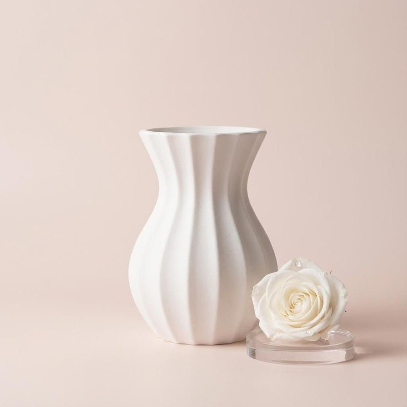 White matte textured fluted minimalist vase beside a cream rose on a clear round stand, against a soft blush pink background.