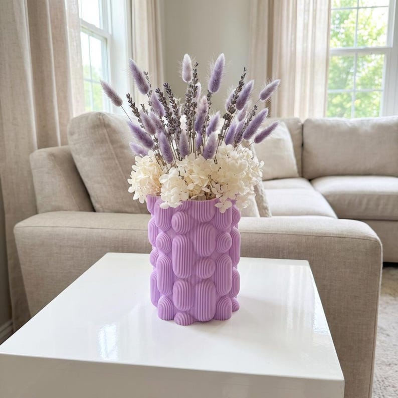 Textured matte lavender unique vase with lavender and white flowers on a glossy white table in front of a beige sofa.