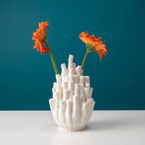 Matte white coral vase with orange flowers in its tubes, set on a white surface against a deep teal background.
