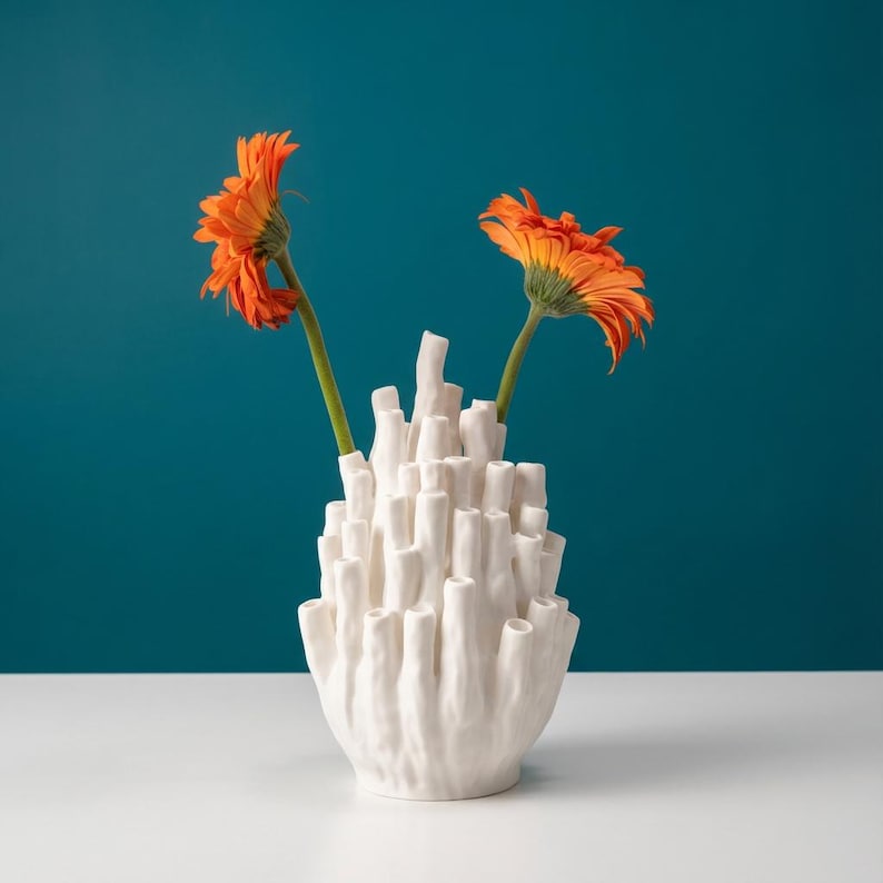 Matte white coral vase with orange flowers in its tubes, set on a white surface against a deep teal background.