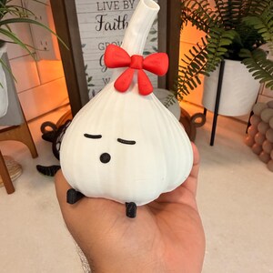 Garlic night light, a cream-white garlic-shaped lamp with a small red bow at the top and a sleepy face, held in an open hand in front of a lit shelf with plants and a wooden sign.