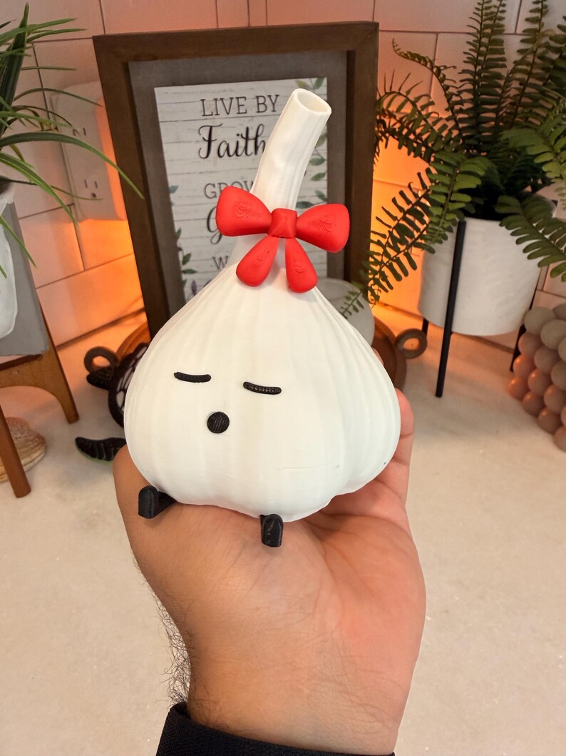 Garlic night light, a cream-white garlic-shaped lamp with a small red bow at the top and a sleepy face, held in an open hand in front of a lit shelf with plants and a wooden sign.