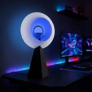 Modern table lamp with a pleated white ring shade glowing blue, on a matte black pyramid base on a dark gaming desk with a monitor and pink and purple accent lights.