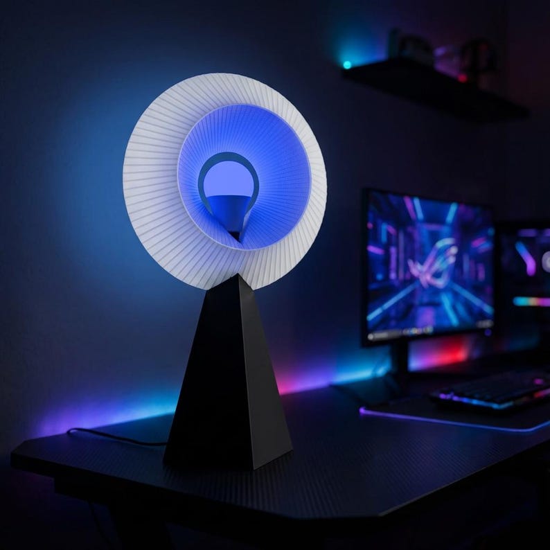 Modern table lamp with a pleated white ring shade glowing blue, on a matte black pyramid base on a dark gaming desk with a monitor and pink and purple accent lights.
