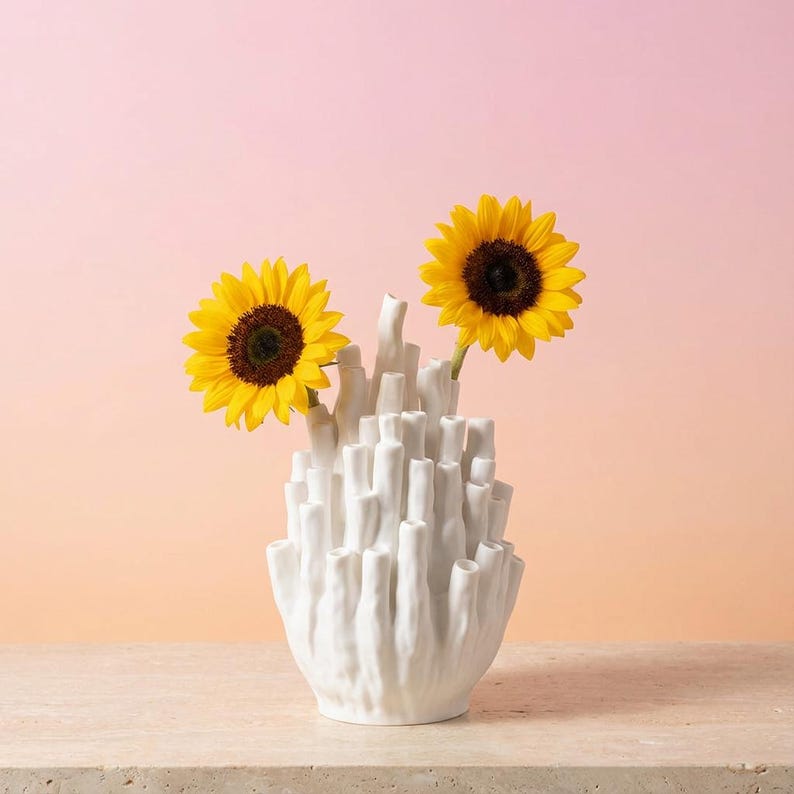 Matte white textured coral vase holding yellow sunflowers against a warm pink and peach background.