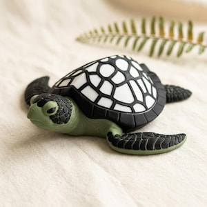 May include: A small sea turtle figurine with a sage green head and flippers featuring black textured scales. The shell is white with a bold black geometric pattern. The turtle is positioned in a forward-facing orientation with its head slightly turned.