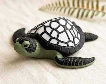 May include: A small sea turtle figurine with a sage green head and flippers featuring black textured scales. The shell is white with a bold black geometric pattern. The turtle is positioned in a forward-facing orientation with its head slightly turned.