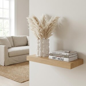 Small textured matte white vase with dried pampas grass on a light wood floating shelf with books, above a living room sofa.