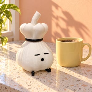 May include: A white garlic-shaped figurine with a black band around its middle, featuring a simple face with closed eyes and a small circular mouth, accompanied by a yellow ceramic mug filled with a dark brown liquid beverage.