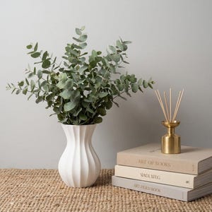 White matte textured fluted modern vase with eucalyptus stems on a woven mat beside a stack of books and a reed diffuser with a gold cap, against a light gray wall.