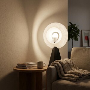 Small table lamp with a pleated matte white ring shade glowing warm white, on a matte black base on a round wooden side table beside a cream sofa with a knit blanket and a mug.