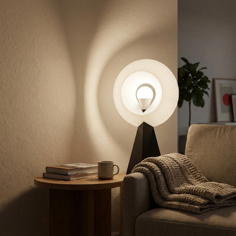 Small table lamp with a pleated matte white ring shade glowing warm white, on a matte black base on a round wooden side table beside a cream sofa with a knit blanket and a mug.