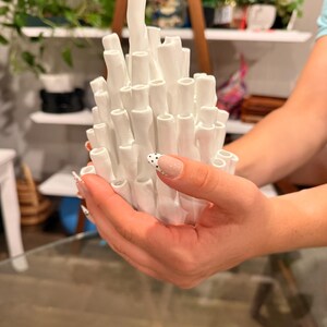 Hand holding a white coral ikebana vase with many tall tubular stems, matte white texture, held up in front of shelves with green plants, showing its compact palm-sized scale.