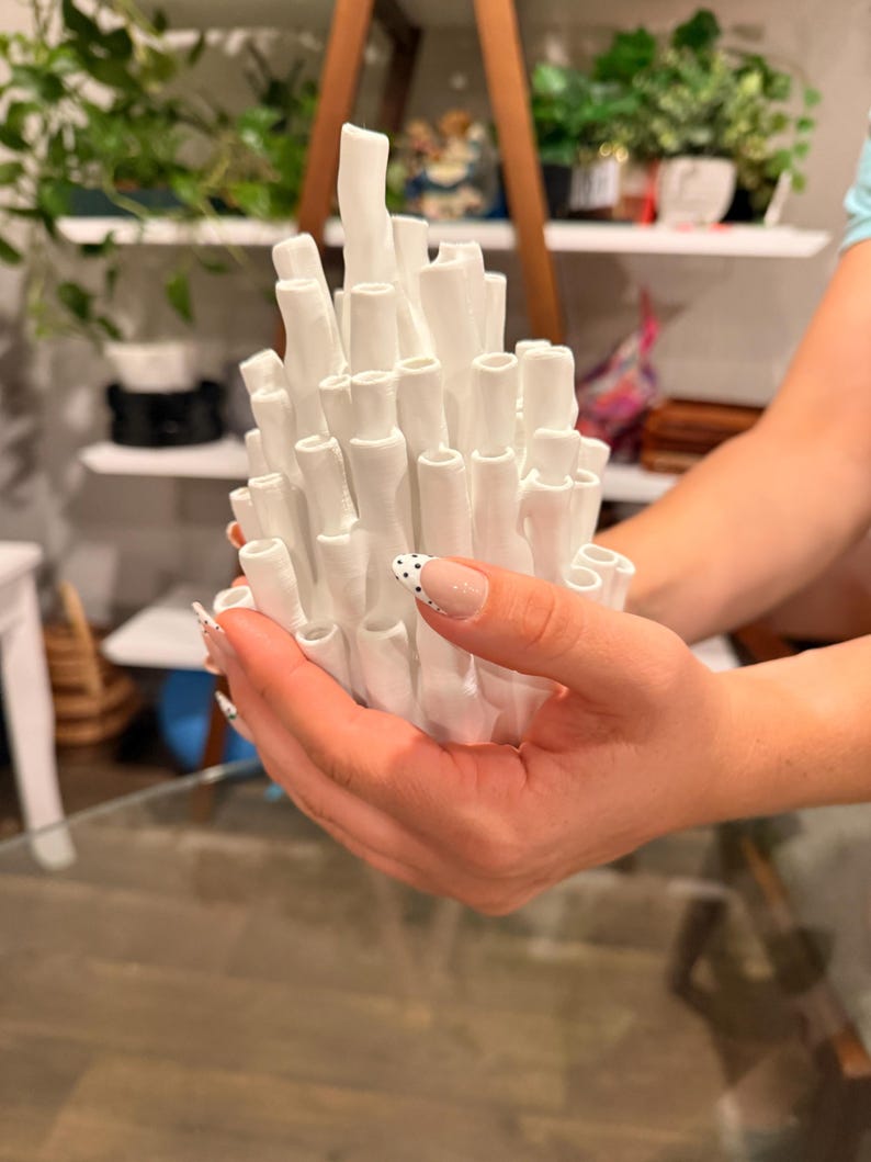 Hand holding a white coral ikebana vase with many tall tubular stems, matte white texture, held up in front of shelves with green plants, showing its compact palm-sized scale.