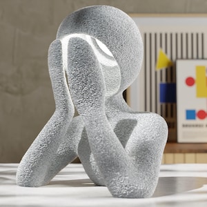 以下が含まれることがあります： 顔を覆う手をした人の薄い灰色の彫刻。彫刻はテクスチャ加工された表面を持ち、白い表面に置かれています。背景には額入りのアートプリントがあります。