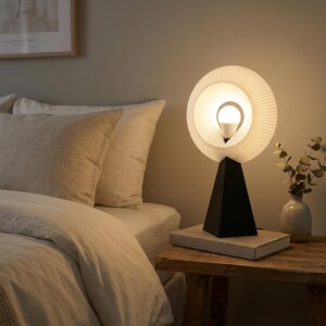 Modern table lamp with a pleated matte white ring shade glowing warm amber, on a matte black base on a book on a wooden nightstand beside a bed and a small vase of dried stems.