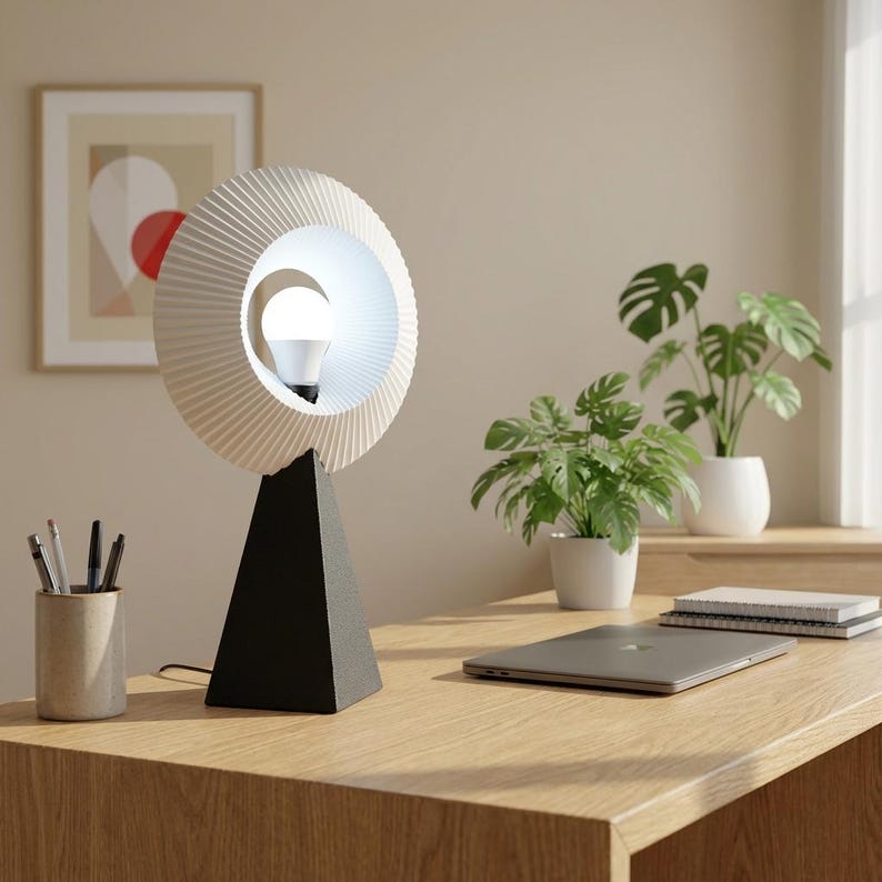Modern table lamp with a round pleated matte white ring shade glowing soft white around a bulb, on a matte black pyramid base, on a wooden desk beside plants and a laptop. About 14.5 inches tall with a 10 inch shade.