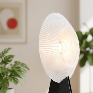 Close-up of the pleated, textured white ring shade of a modern table lamp, glowing warm white around the bulb, above a matte black base with a blurred plant behind.