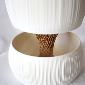 Close-up of a textured matte white ribbed pot nested above its lower bowl, the brown cone-shaped core visible between them, on white fabric; a self watering planter design.