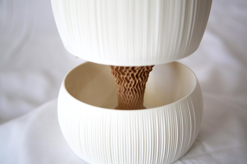 Close-up of a textured matte white ribbed pot nested above its lower bowl, the brown cone-shaped core visible between them, on white fabric; a self watering planter design.