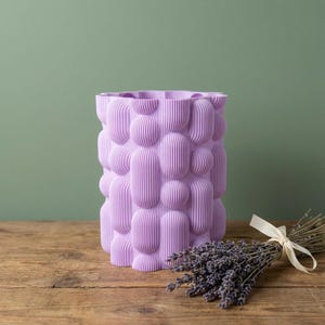 Textured matte lavender small vase on a wooden tabletop with a bundle of dried lavender tied with ribbon against a sage green wall.