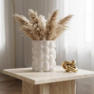 Textured matte white vase with dried pampas grass on a cream-colored pedestal table beside a gold knot ornament, a small vase for soft neutral decor.