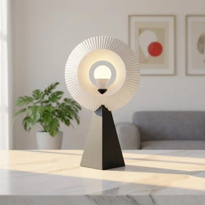 Modern table lamp with a pleated matte white ring shade glowing soft white, on a matte black pyramid base on a white-veined tabletop, with framed art, a plant and a gray sofa behind.