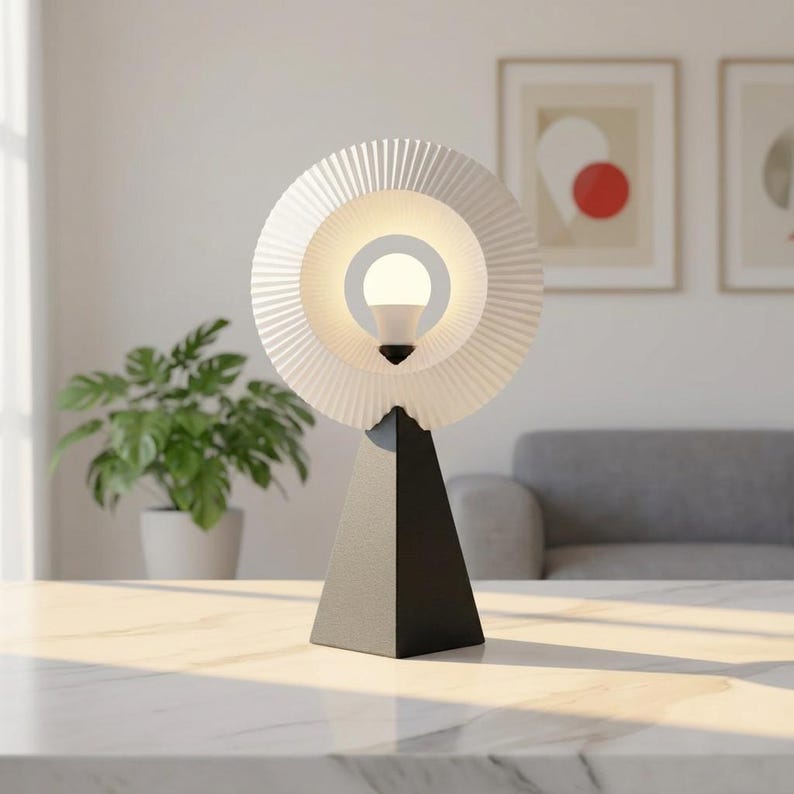 Modern table lamp with a pleated matte white ring shade glowing soft white, on a matte black pyramid base on a white-veined tabletop, with framed art, a plant and a gray sofa behind.