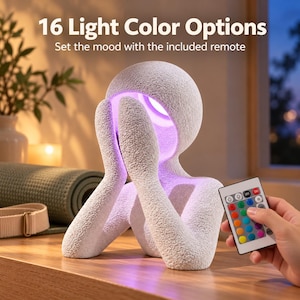 Night light with a matte textured white figure glowing soft purple, held beside a hand pointing a colorful light remote, on a wooden shelf with a window behind and text reading Light Color Options.