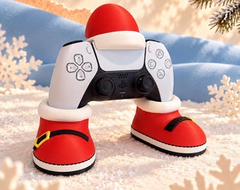 May include: A white and black video game controller resting inside a red and white Santa-themed stand. The stand features two red boots with white fur trim and black straps, and a red Santa hat sits on top of the controller.